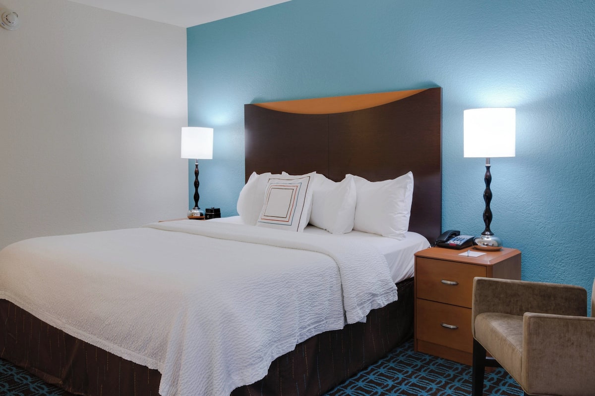 Une chambre d’hôtel avec un grand lit, deux tables de chevet, une chaise et une petite table de nuit avec une lampe.