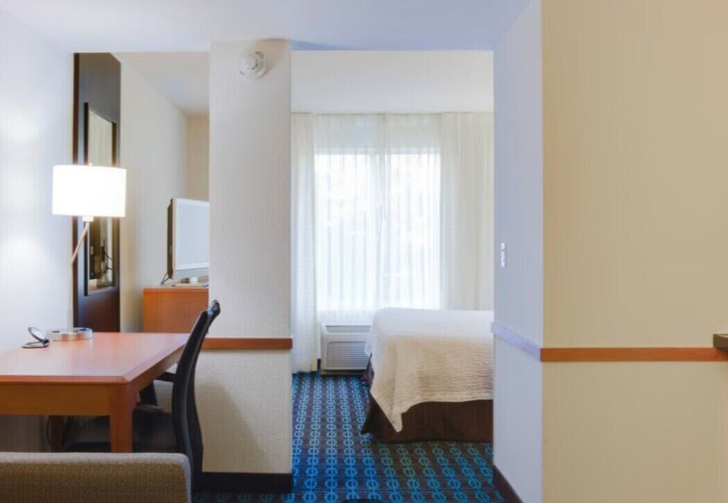 A hotel room with a bed, a desk with a lamp, a chair, a window with curtains, and a patterned carpet.