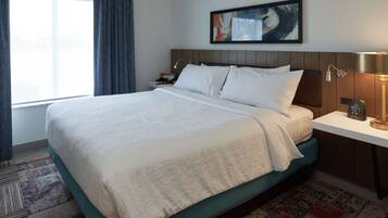 A hotel room with a large bed, a bedside table with a lamp, a nightstand, a wall-mounted artwork, and a window with curtains.