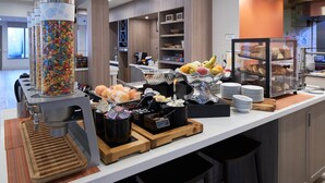 Zona de bufé con diversos alimentos, incluyendo una máquina dispensadora de dulces, frutas y pasteles.