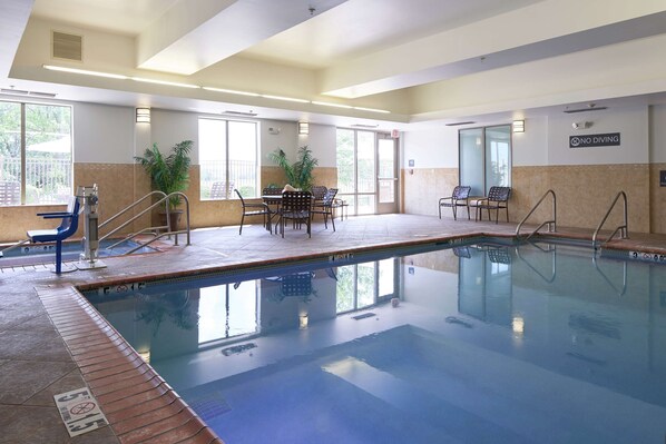 Une piscine intérieure à l’eau claire, entourée de chaises et de tables, et un panneau indiquant « Il est interdit de plonger » est affiché sur le mur.
