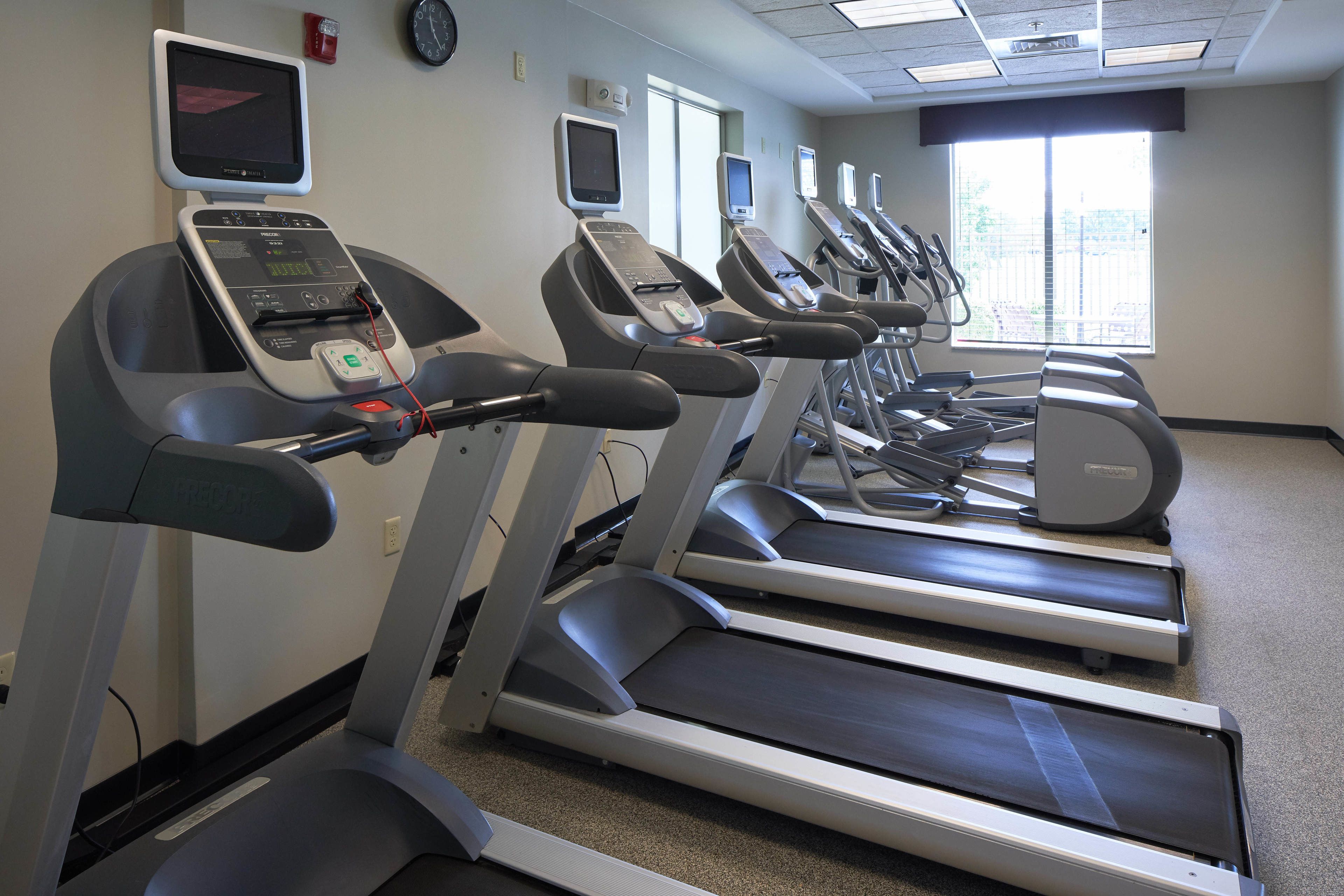 A row of treadmills in a gym with digital displays, a clock on the wall, and a window with blinds.