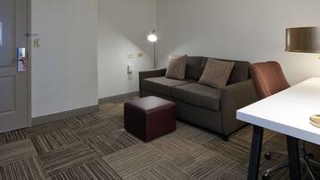 A hotel room with a grey sofa, a small ottoman, a white desk, and a floor lamp.