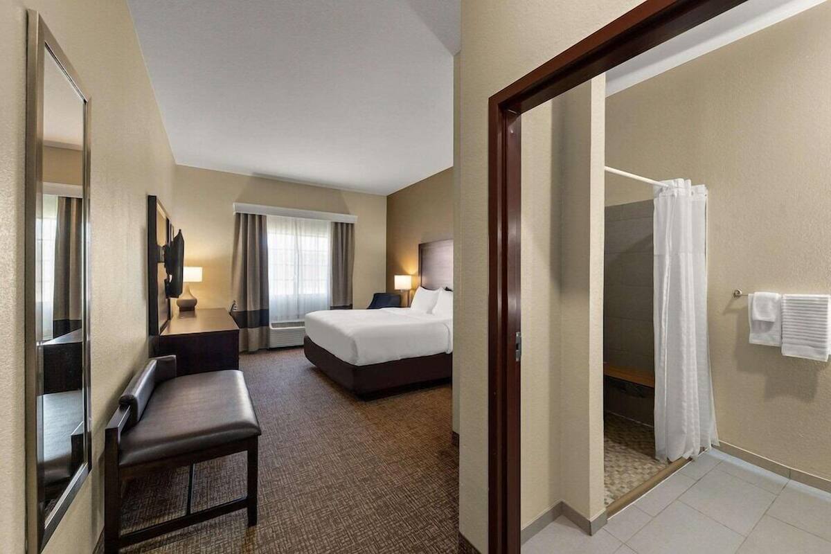 A hotel room with a bed, a desk, a chair, a mirror, a shower, and a bathroom with a towel rack.