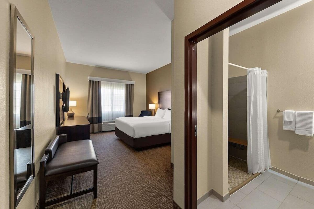 A hotel room with a bed, a desk, a chair, a mirror, a shower, and a bathroom with a towel rack.