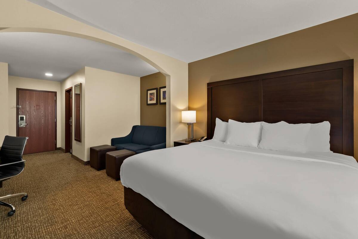 A hotel room with a large bed, a desk with a chair, a sofa, a lamp, and a door.