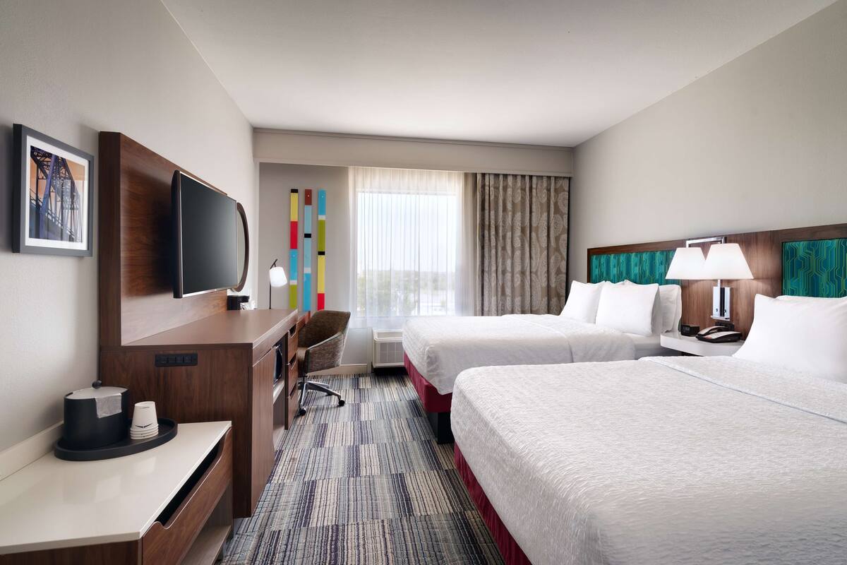 A hotel room with two beds, a desk with a TV, a chair, a window with curtains, and a wall-mounted artwork.