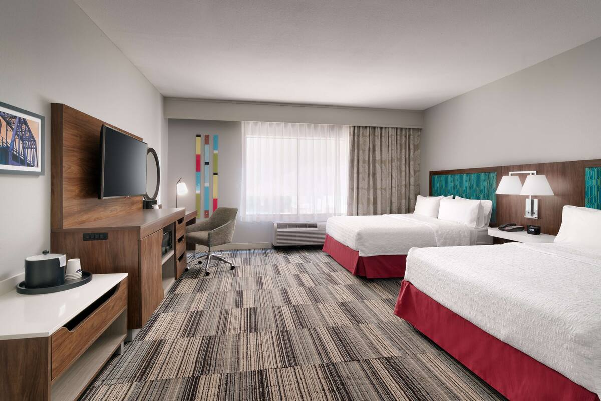 A hotel room with two beds, a desk with a TV, a chair, a desk lamp, a bedside table with a lamp, and a large window with curtains.