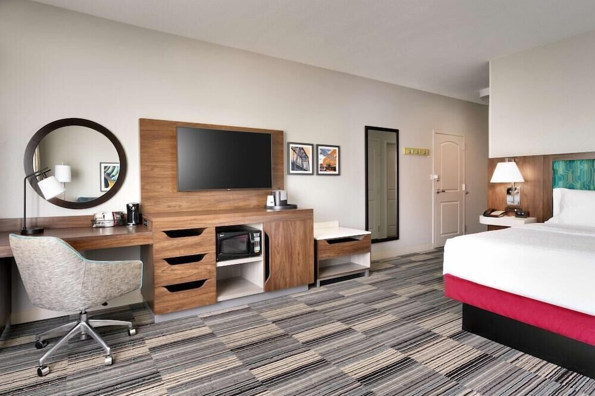 A hotel room with a bed, a desk with a chair, a TV, a microwave, and a door.