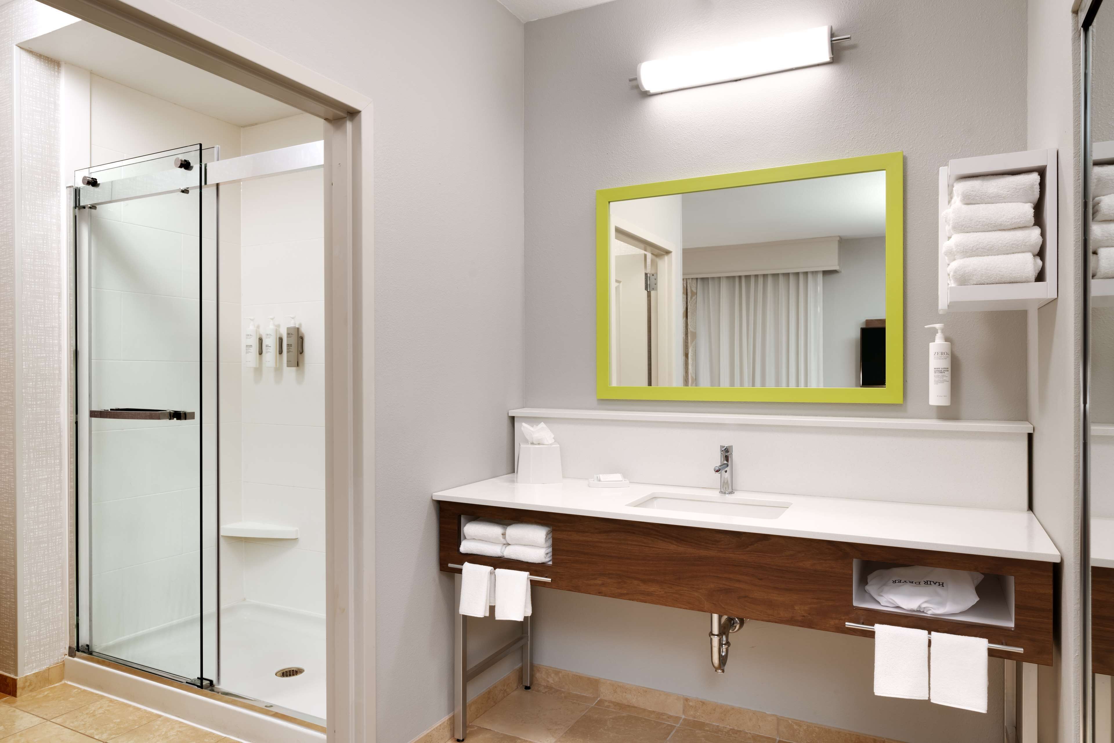 A modern bathroom with a glass shower enclosure, a white sink with a wooden vanity, a mirror with a green frame, and neatly arranged towels.