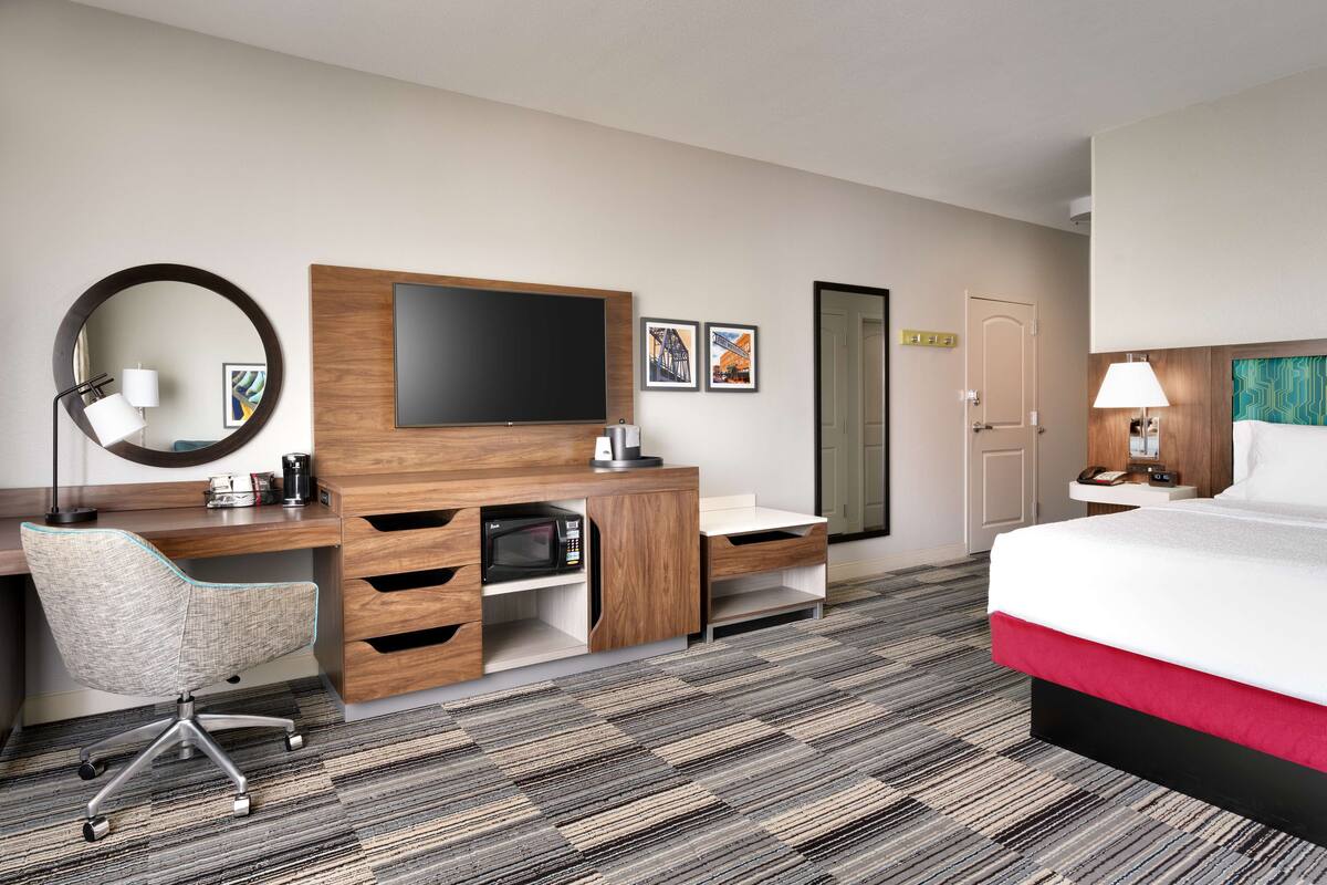 A hotel room with a bed, a desk with a chair, a TV, a microwave, and a door.