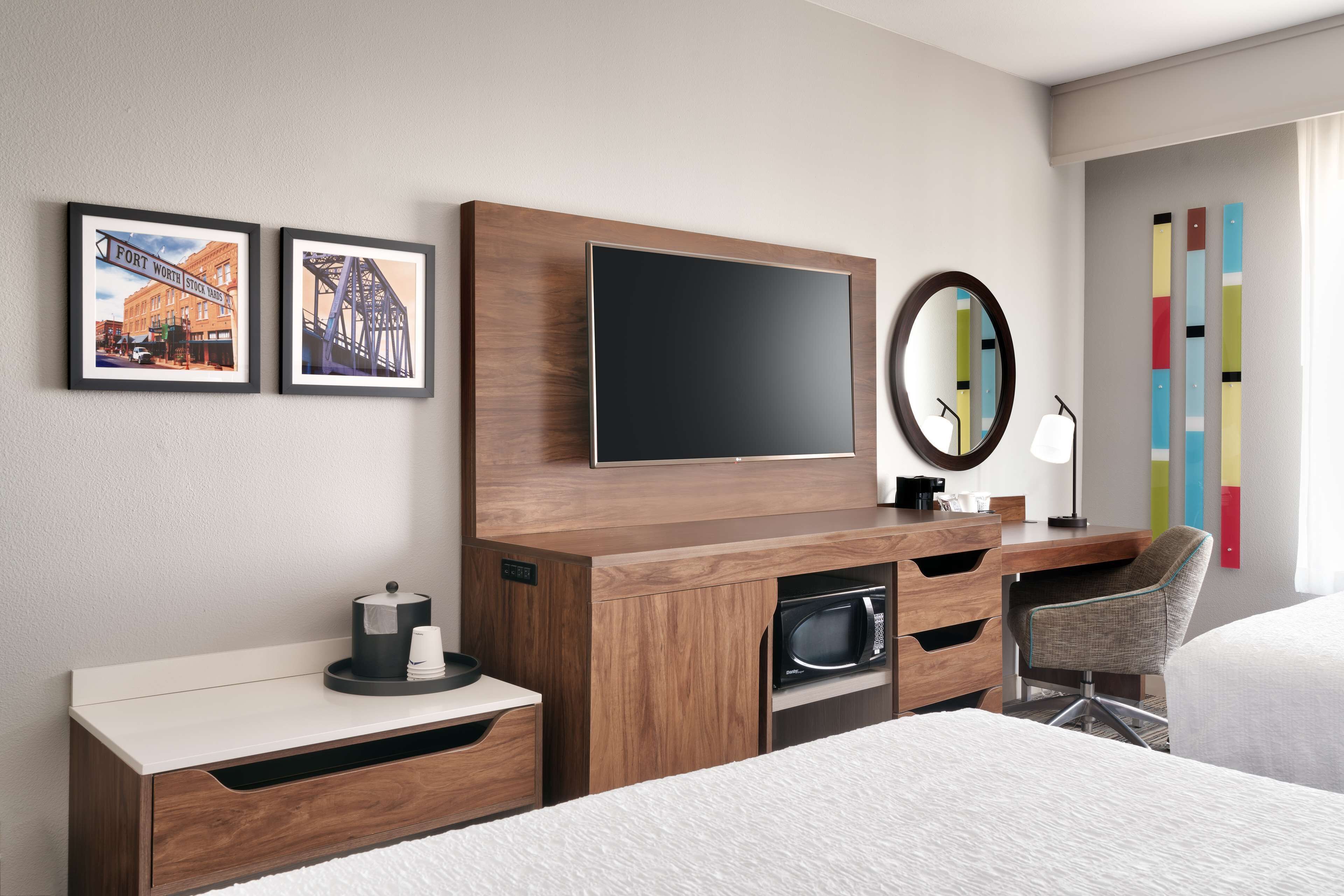 A hotel room with a wooden desk, a flat-screen TV, a microwave, a chair, and two framed pictures on the wall.