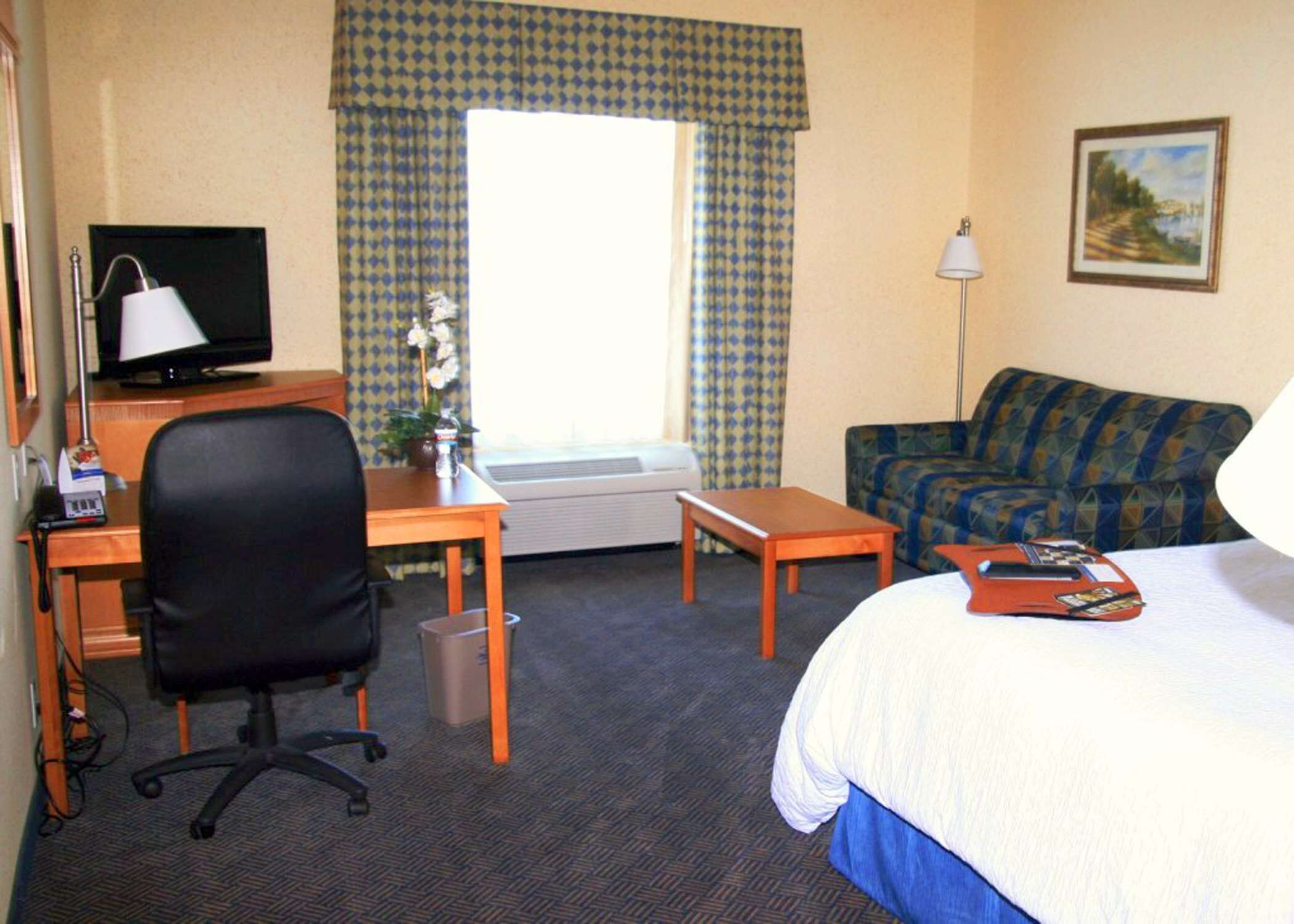 Une chambre d’hôtel comprenant un lit, un bureau avec un ordinateur, une chaise, une petite table, un canapé et une fenêtre avec des rideaux.