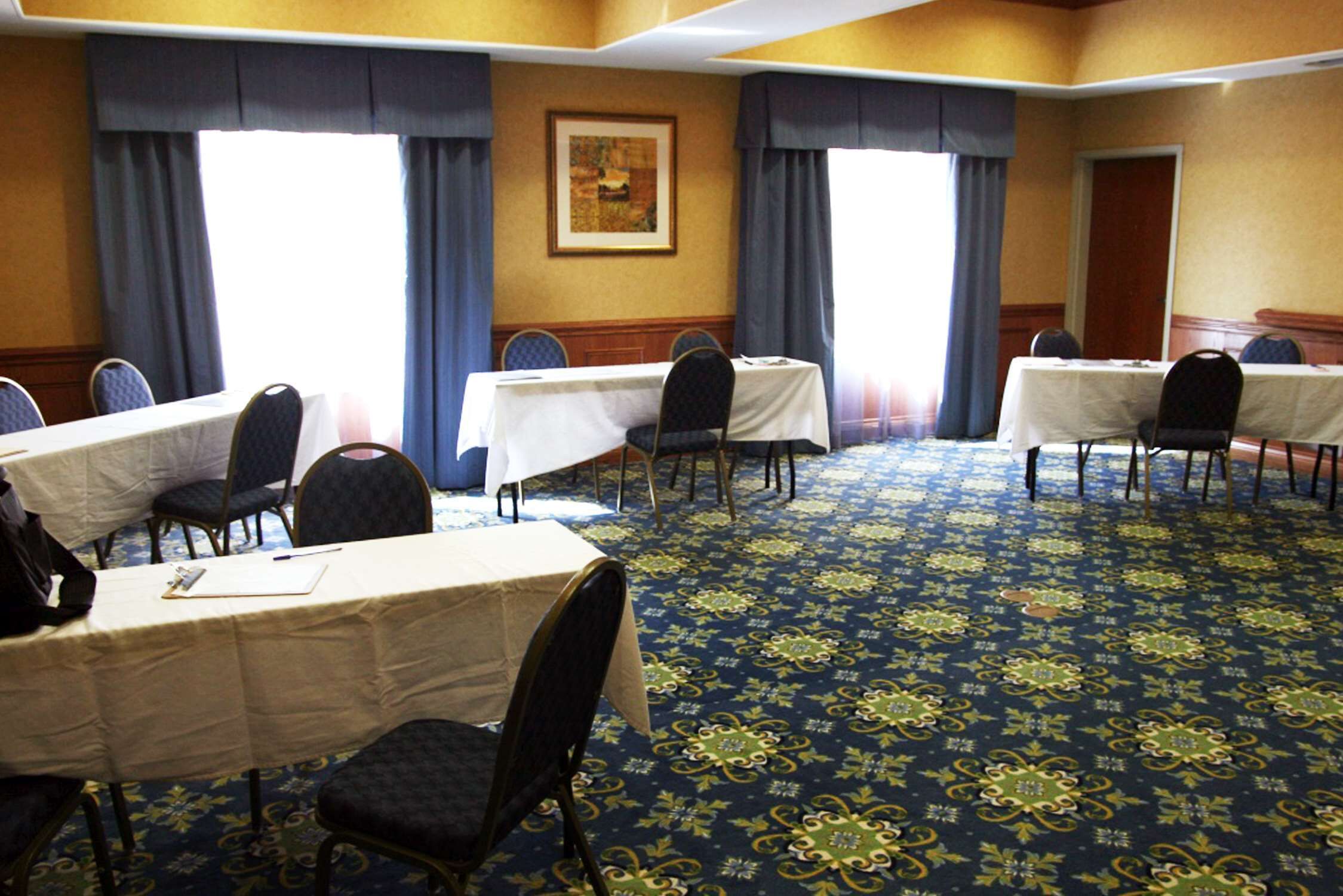 A conference room with multiple tables and chairs, large windows with curtains, and a patterned carpet.