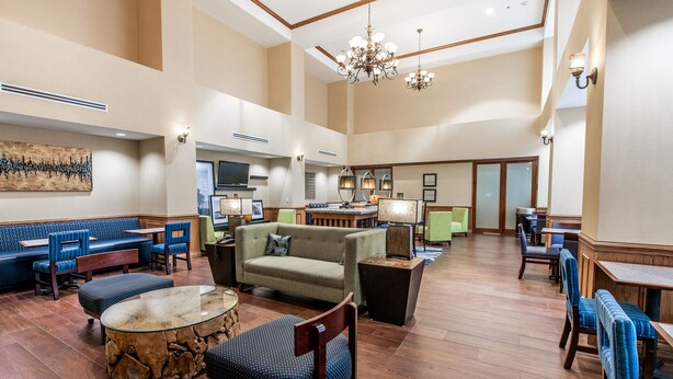 Een ruime lobby met een hoge plafond, houten vloer, een centraal zitgedeelte met banken en stoelen, en een eetgedeelte met tafels en stoelen.
