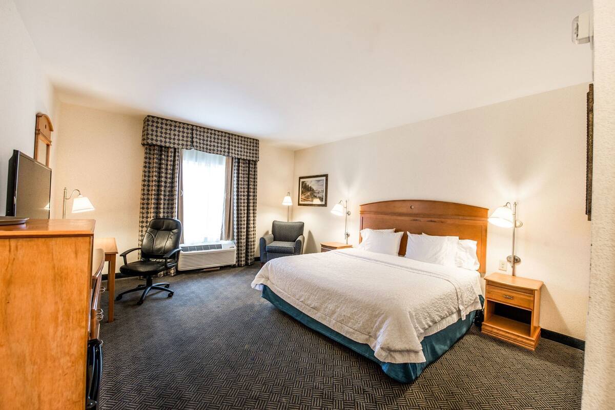 A hotel room with a bed, a desk, two chairs, a TV, and a window with curtains.