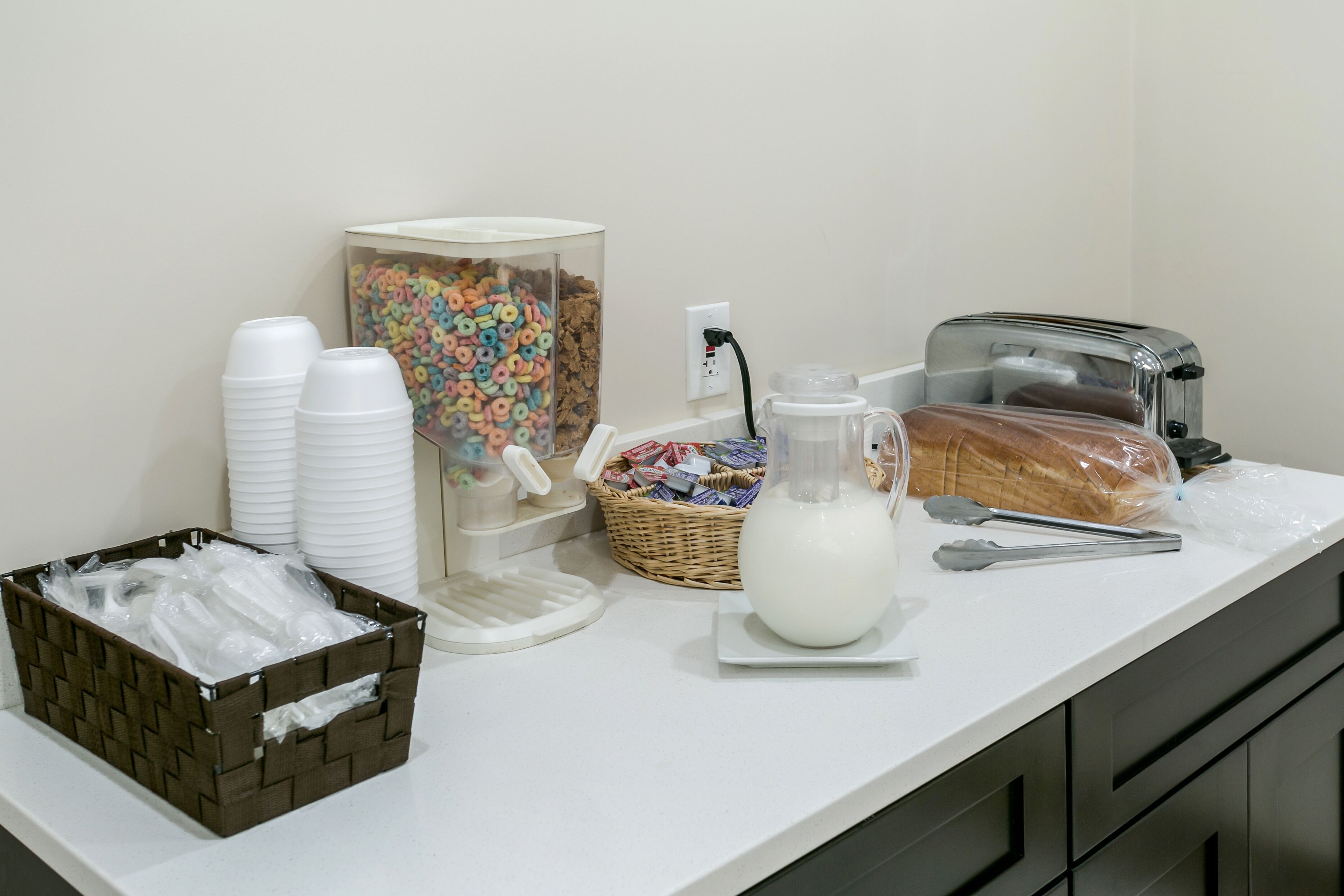 Una disposición para el desayuno con vasos desechables, cereales, una jarra de leche, un tostador y una cesta de bolsas de plástico.