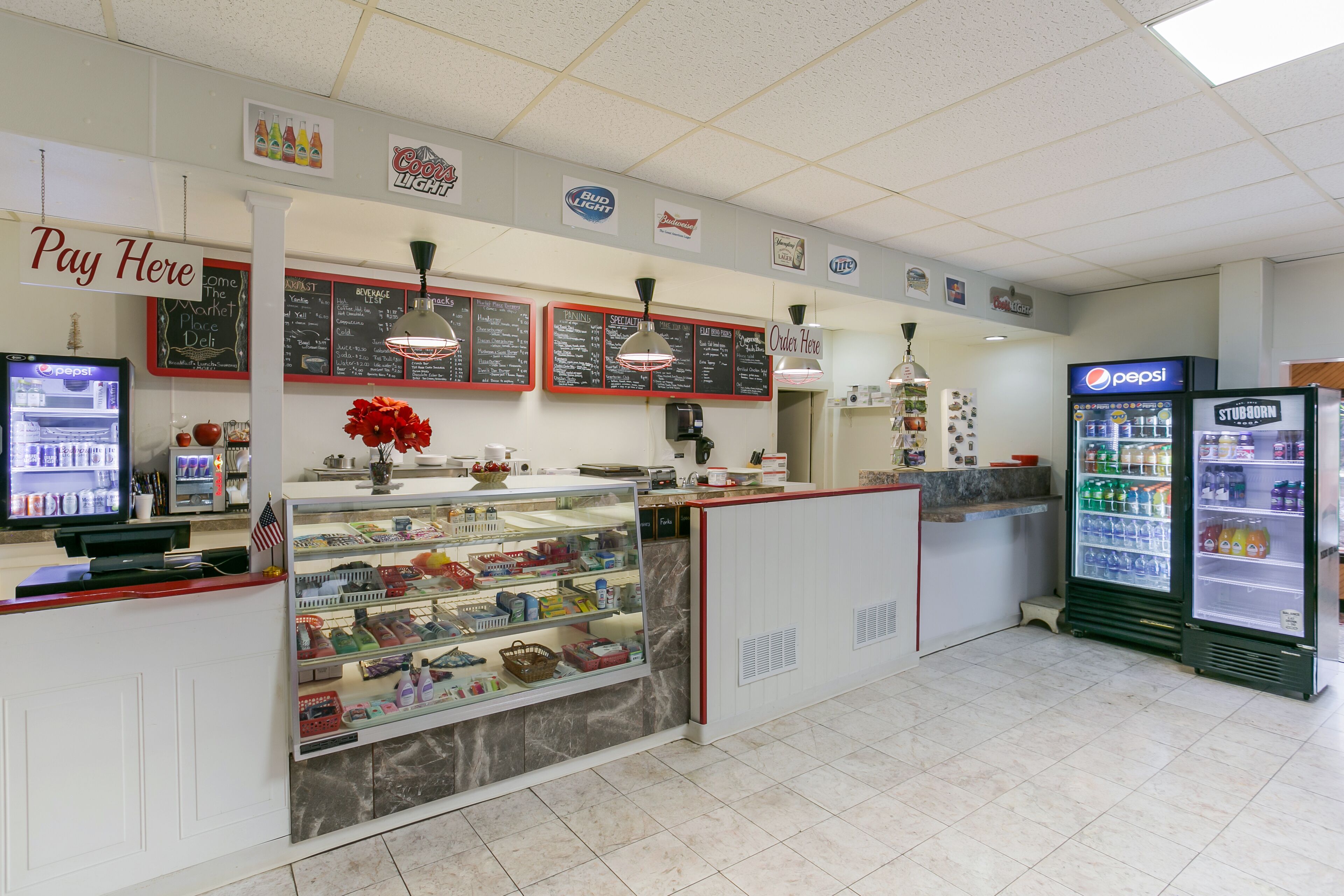 A clean, well-lit convenience store with a 'Pay Here' sign, a Pepsi refrigerator, and various food items on display.