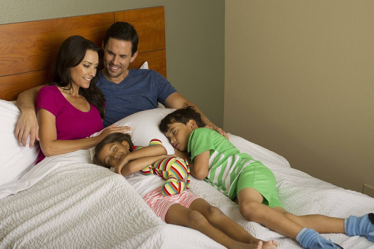 A family of three sleeping in a bedroom with a wooden headboard, white bedding, and a plain wall.