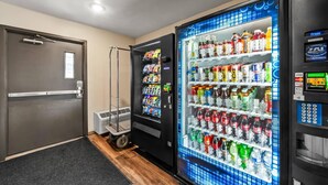 Zona de máquinas expendedoras con un refrigerador lleno de bebidas y aperitivos, un carrito y una puerta.