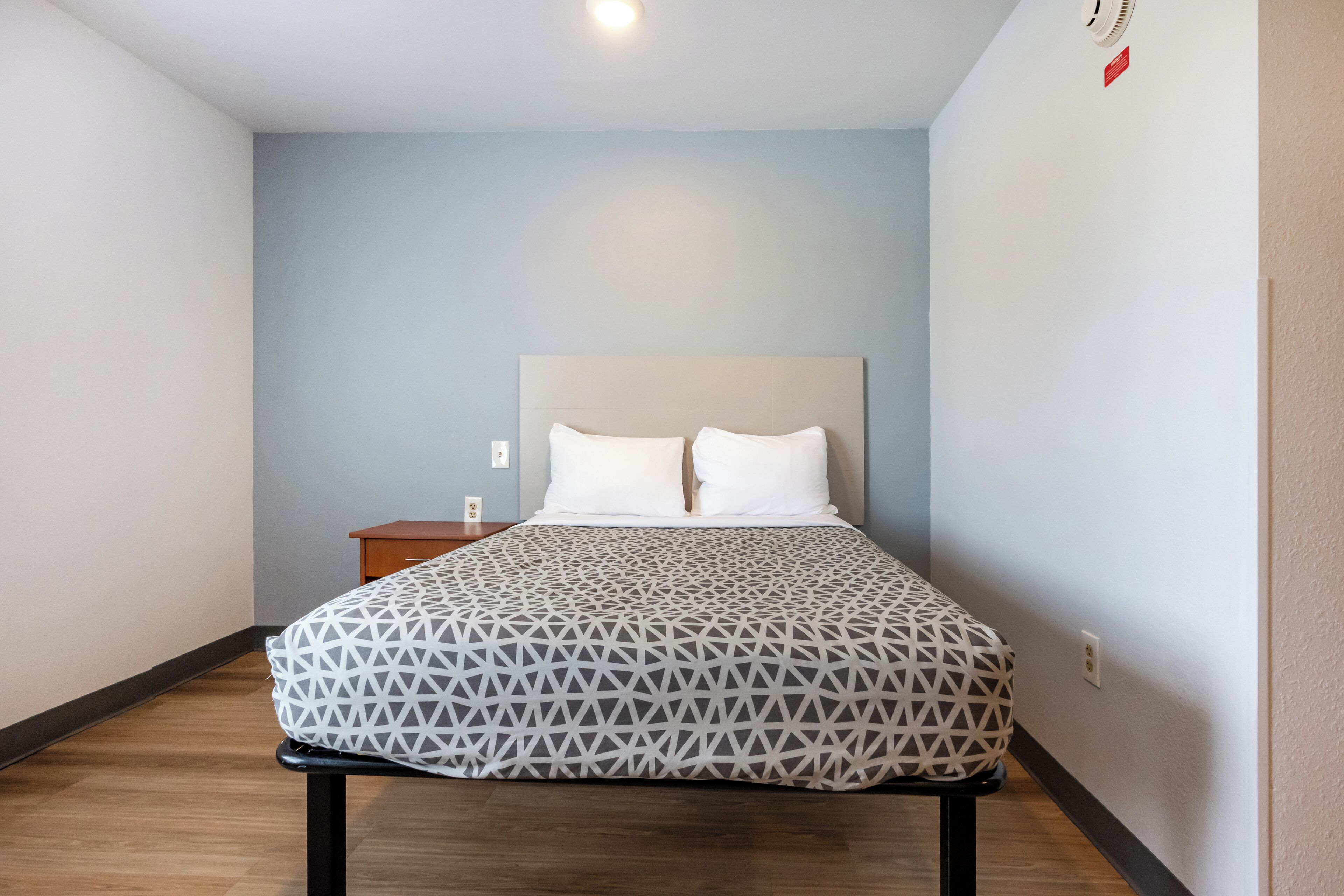 A neatly made bed with a geometric-patterned bedspread, a wooden nightstand, and a wall-mounted light fixture.