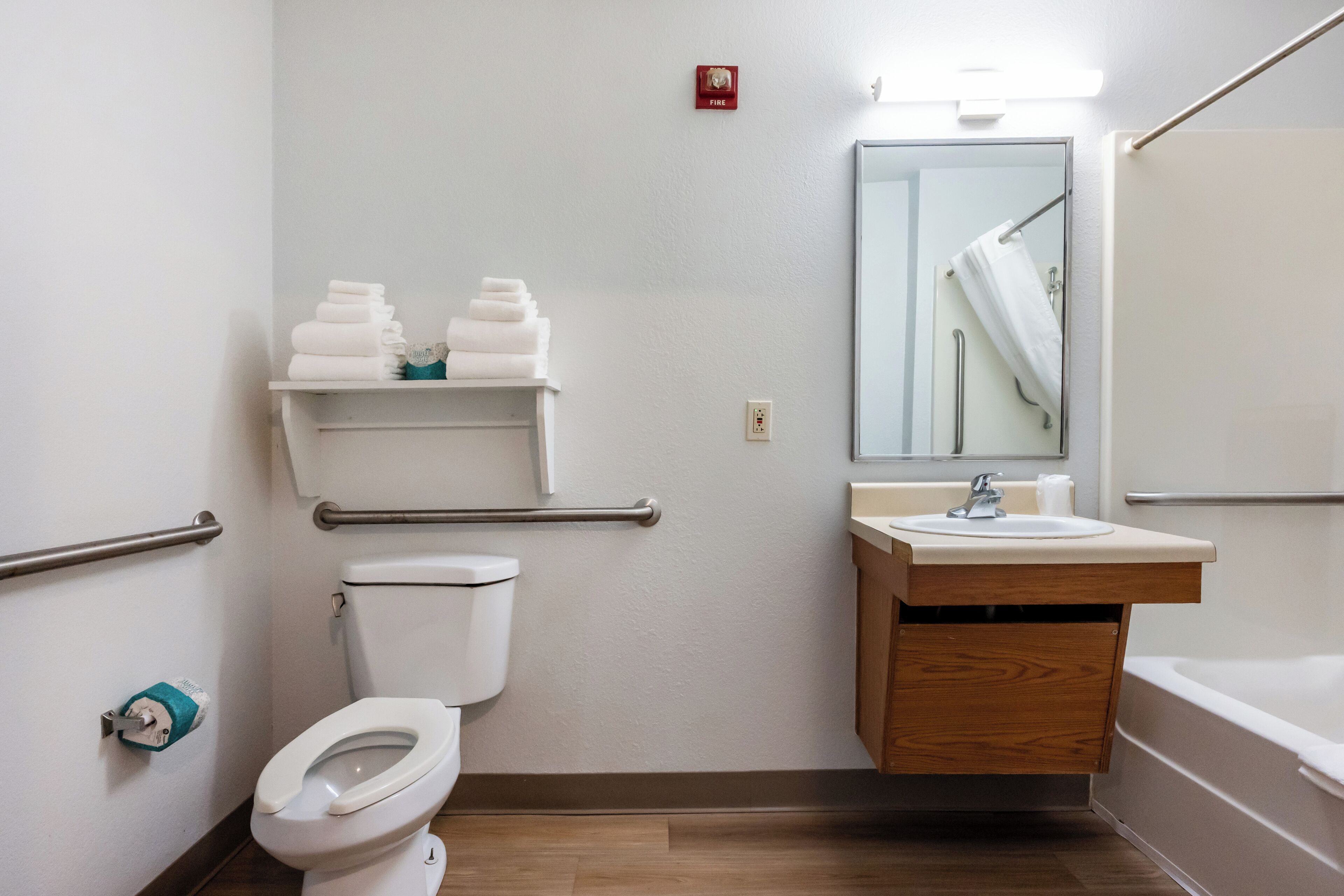 A bathroom with a toilet, sink, and bathtub. There are handrails, a fire alarm, and a mirror above the sink.