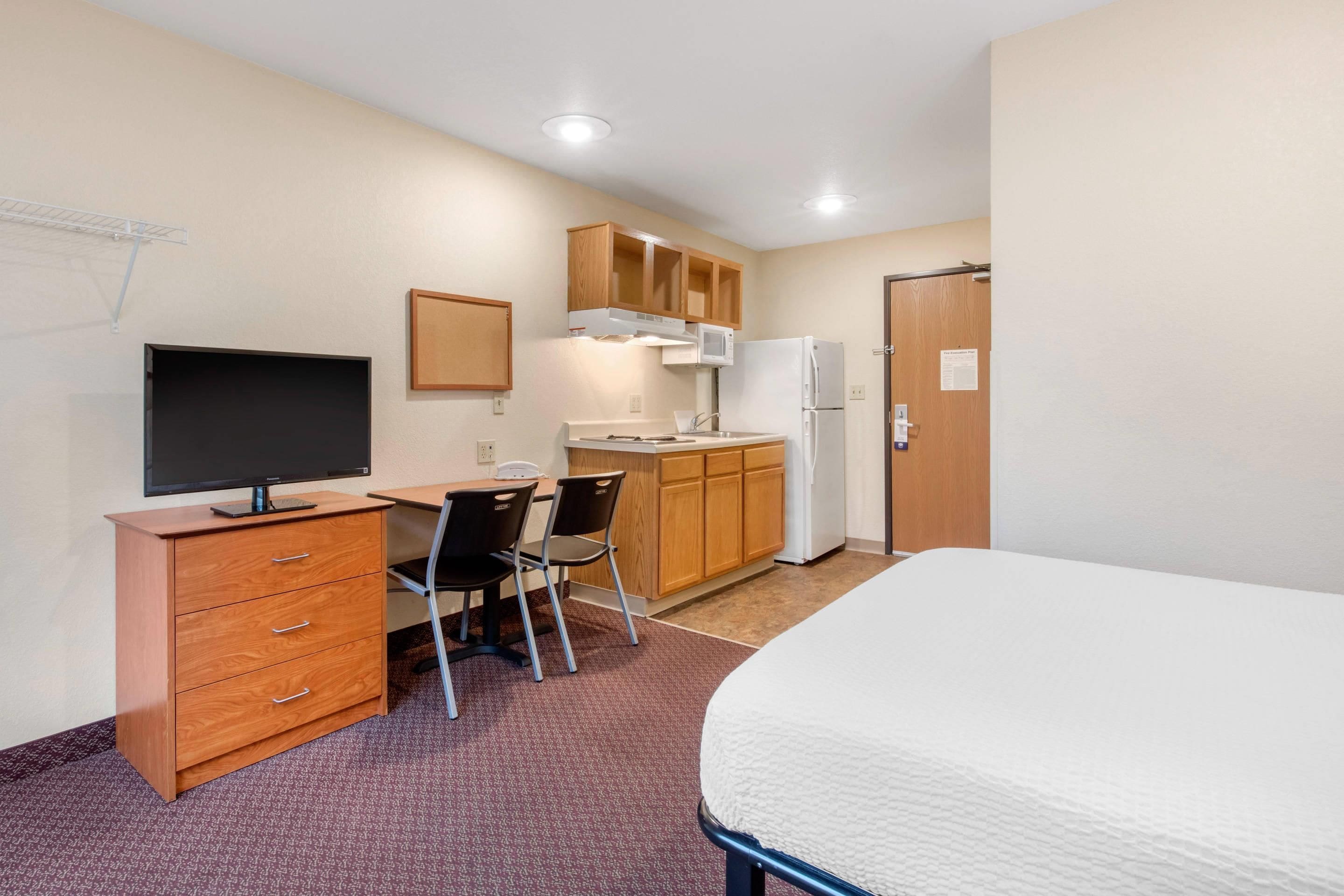 A hotel room with a bed, a desk with a computer, a kitchenette, and a door.