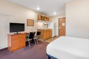 A hotel room with a bed, a desk with a computer, a kitchenette, and a door.