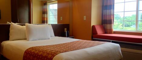 A hotel room with a large bed, a red chair, a window with curtains, and a mirror above the bed.