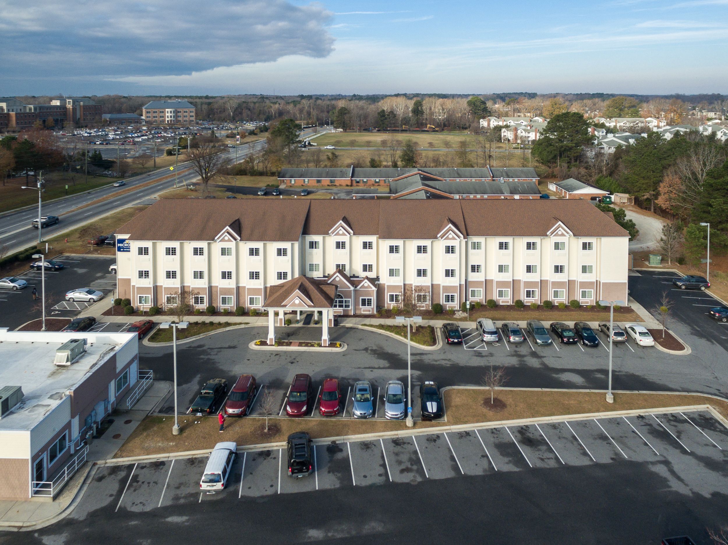 A multi-story hotel with a central entrance, parking lot, and surrounding buildings.