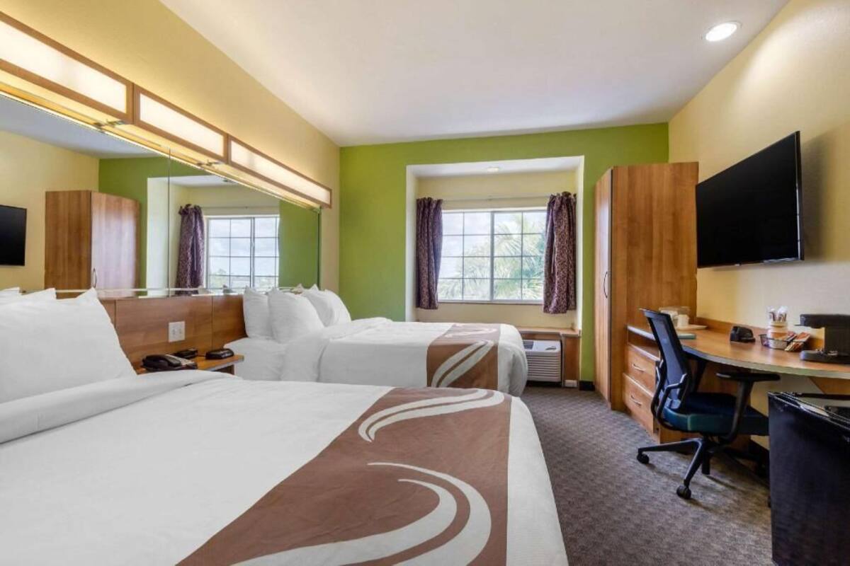 A hotel room with two beds, a desk, a chair, a TV, a window with curtains, and a mirror.