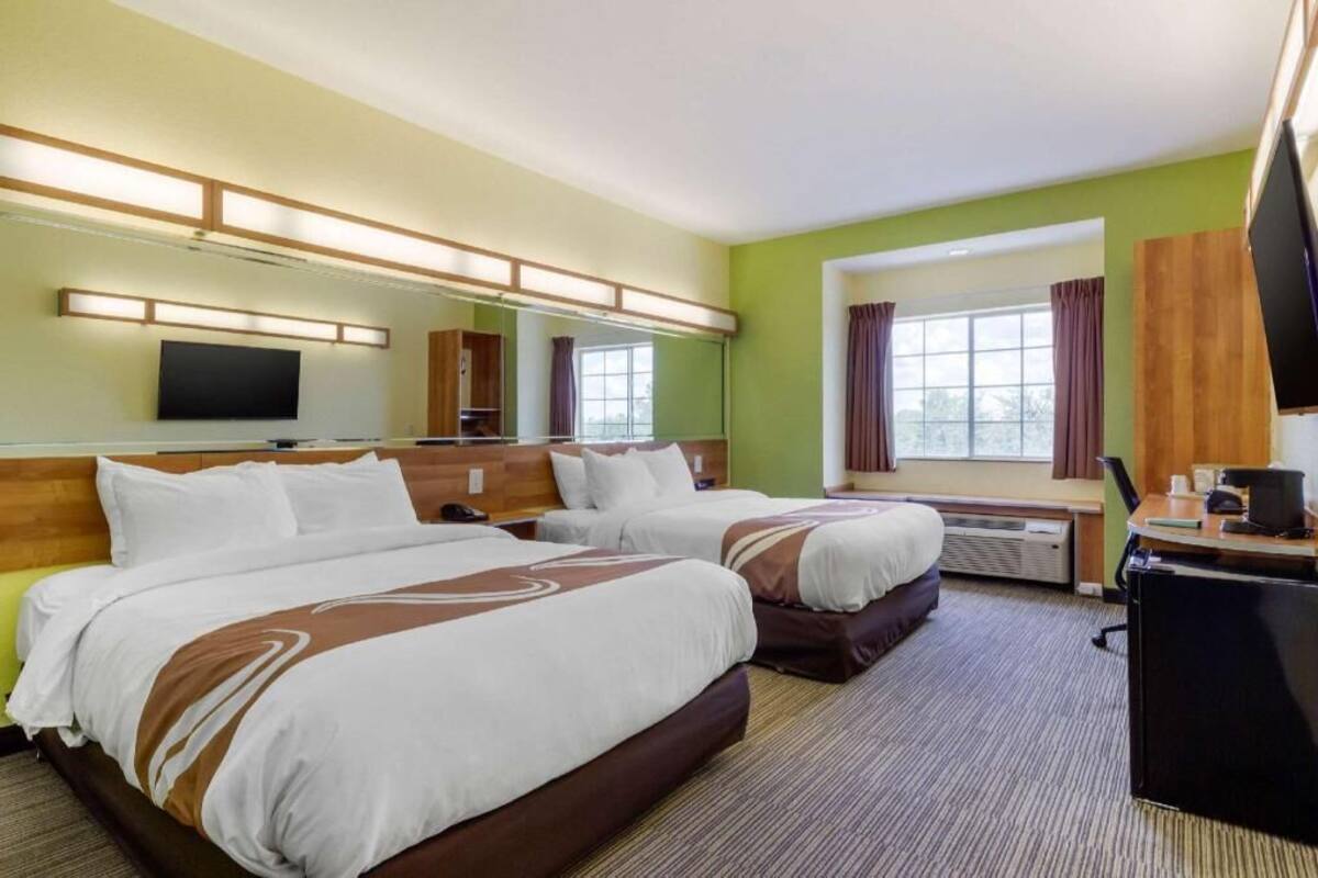 A hotel room with two beds, a desk, a flat-screen TV, a mirror, and a window with curtains.