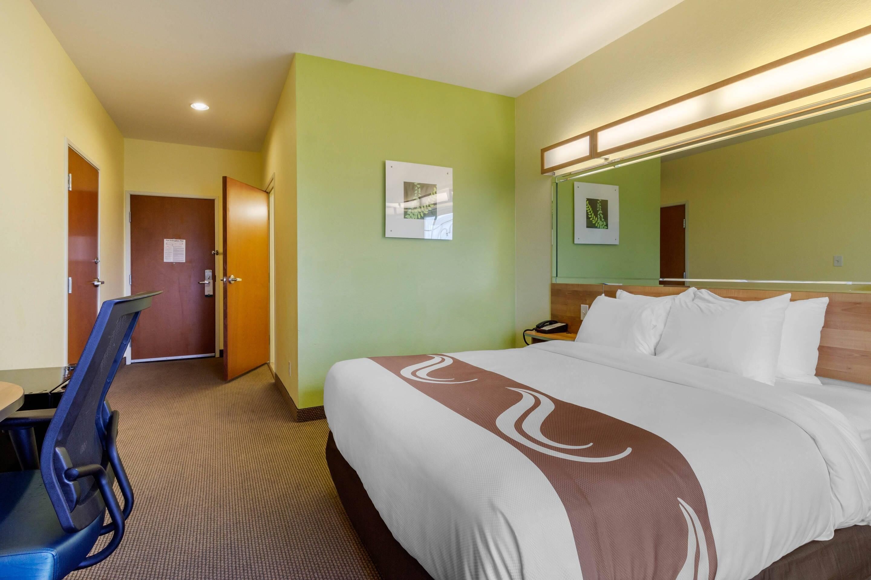 Habitación de hotel con cama, escritorio y silla. Se ve una puerta y un teléfono en la pared.