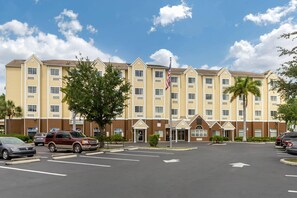 A multi-story hotel with a parking lot in front, featuring a flagpole and several palm trees.