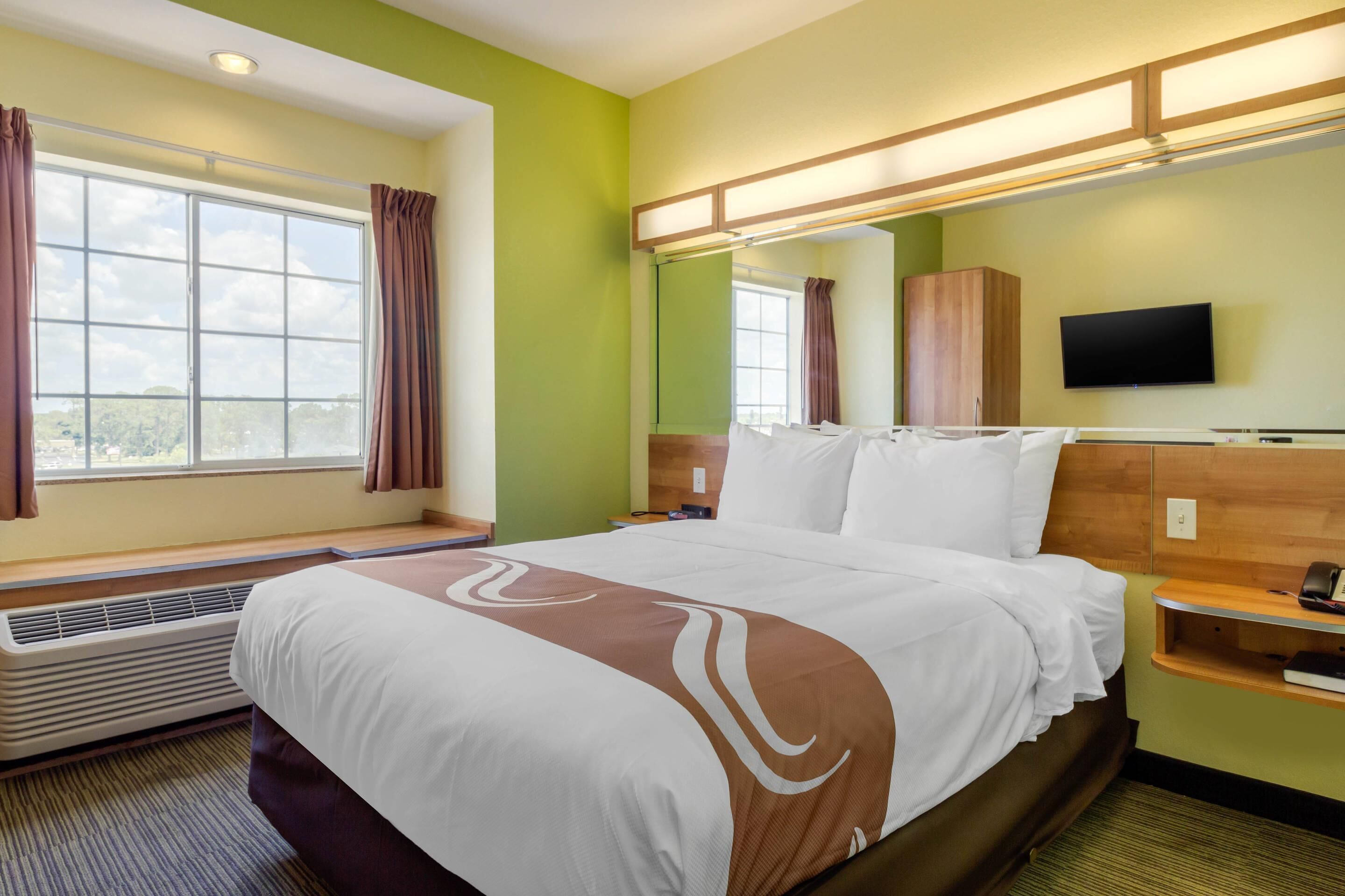 A hotel room with a large bed, a flat-screen TV, a wooden headboard, a nightstand, and a window with curtains.