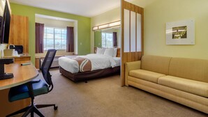 A hotel room with a bed, a desk with a chair, a sofa, a large window with curtains, and a framed picture on the wall. - Quality Inn & Suites Lehigh Acres Fort Myers (Lehigh Acres)