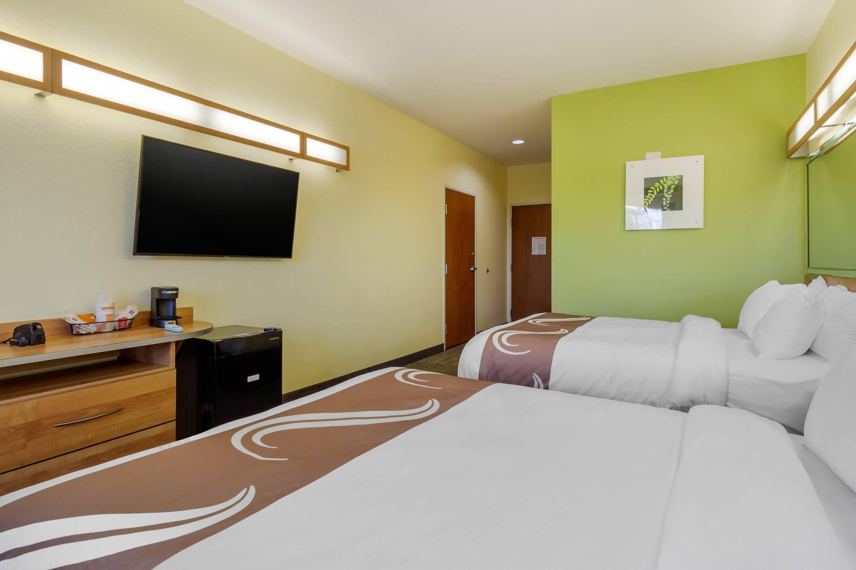 A hotel room with two beds, a desk with a coffee maker, a TV mounted on the wall, and a green accent wall.