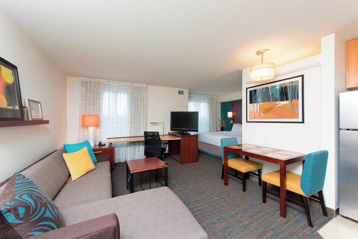 A hotel room with a sofa, a small desk with a chair, a television, a bed, and a dining table with chairs.