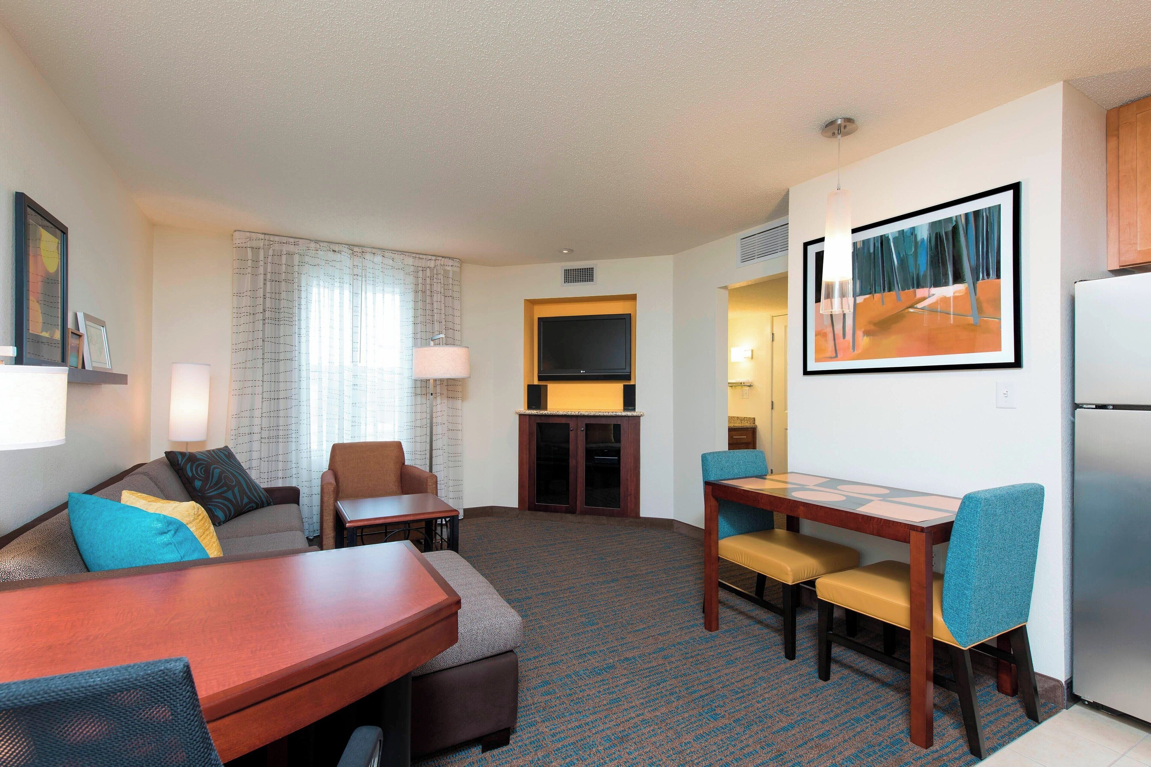 A hotel room with a sofa, a dining table with chairs, a flat-screen TV, a refrigerator, and a painting on the wall.