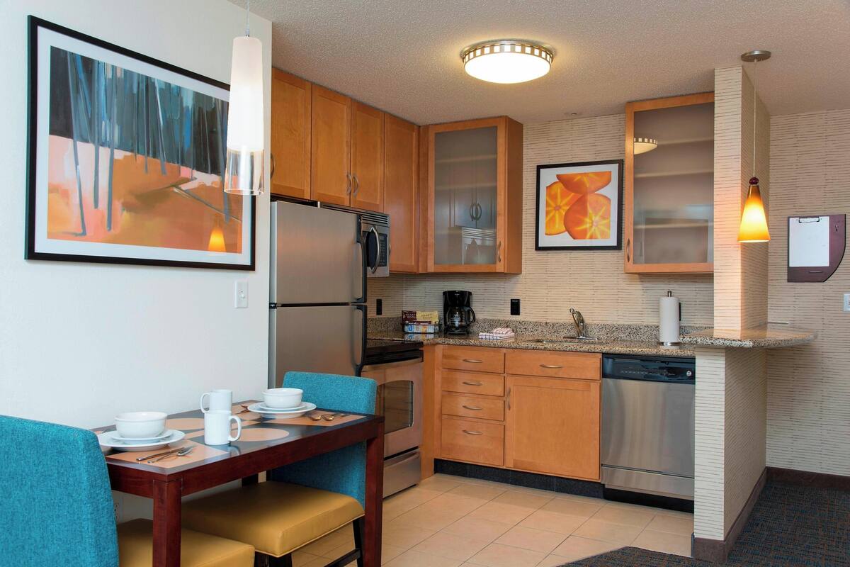 A compact kitchen with wooden cabinets, a built-in dishwasher, and a dining area with a blue chair and a table set for two.