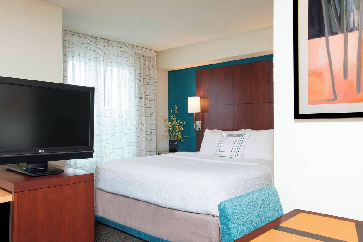 A hotel room with a bed, a desk with a flat-screen TV, a lamp, and a framed picture on the wall.