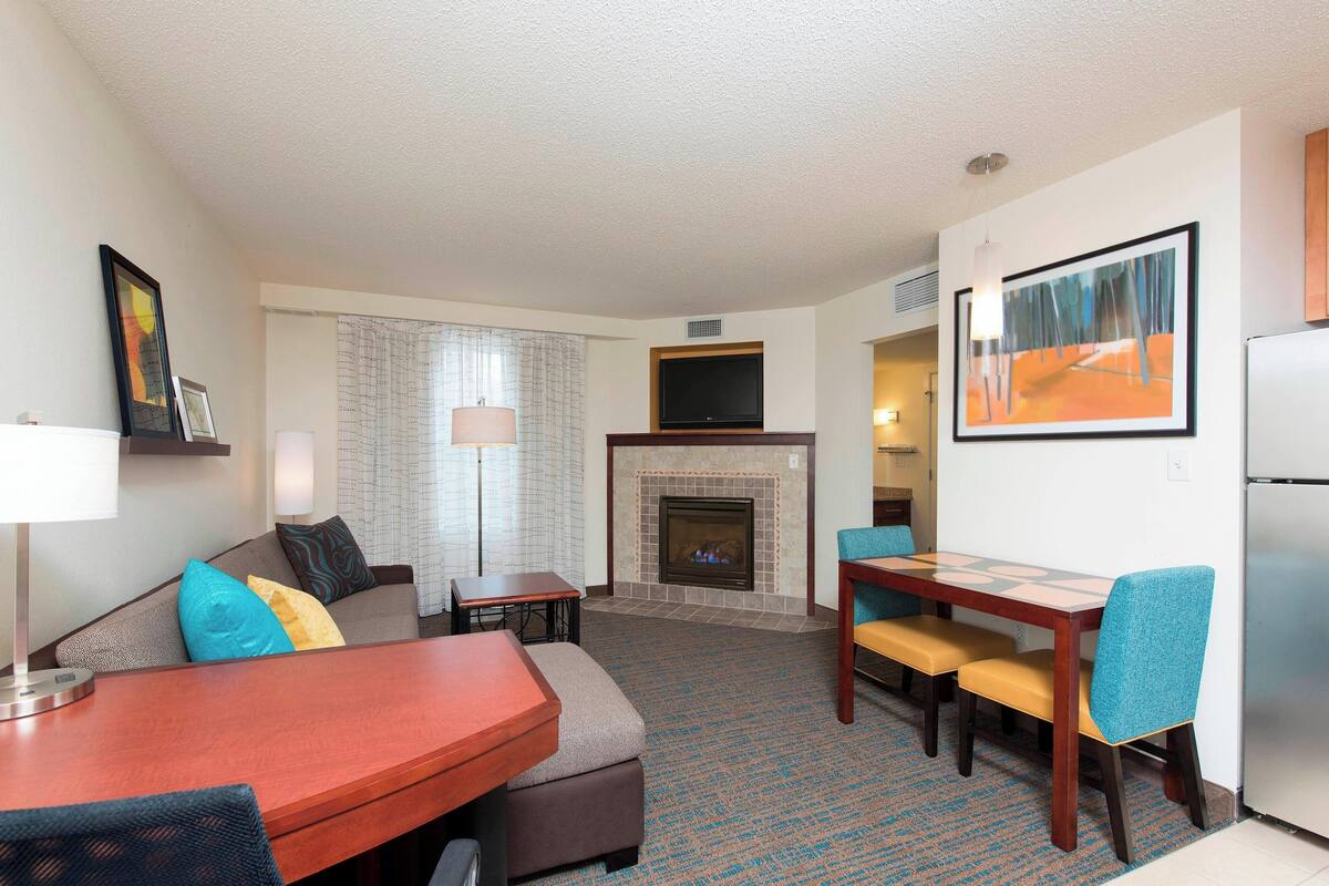 A hotel room with a living area, a fireplace, a dining table with chairs, a TV mounted above the fireplace, and a refrigerator.