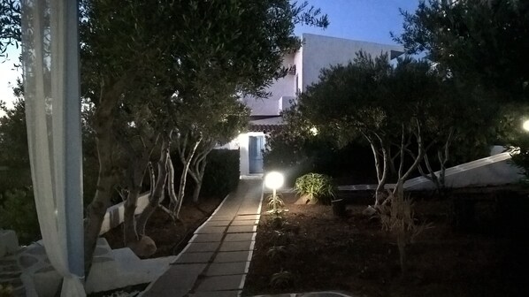 Un chemin menant à un bâtiment à la façade blanche, entouré d’arbres et d’arbustes.