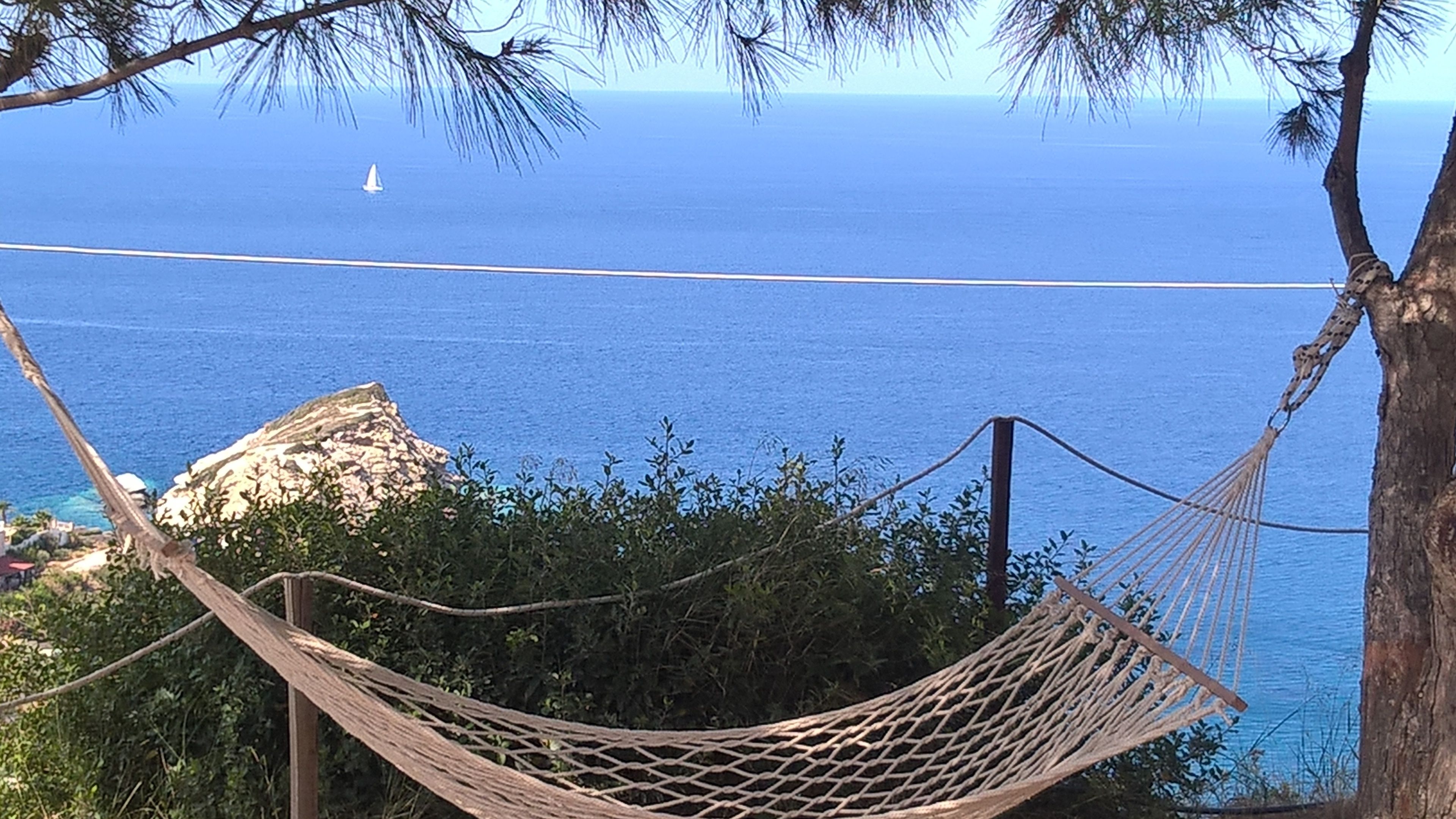 A hammock tied between trees overlooking a clear blue sea with a sailboat in the distance.