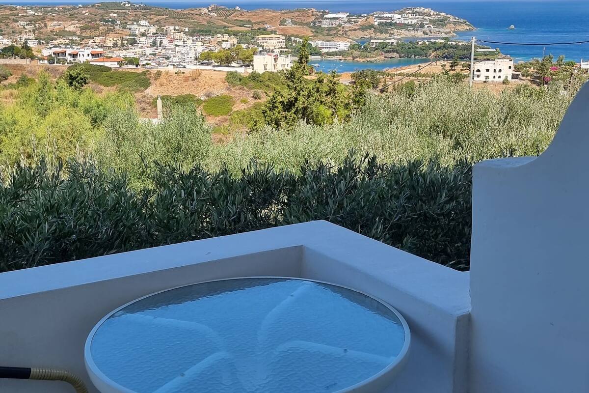 A balcony with a round table and two wicker chairs, overlooking a coastal town and the sea.