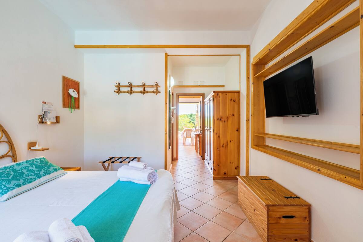 A hotel room with a bed, a TV, wooden furniture, and a view of the outdoors.