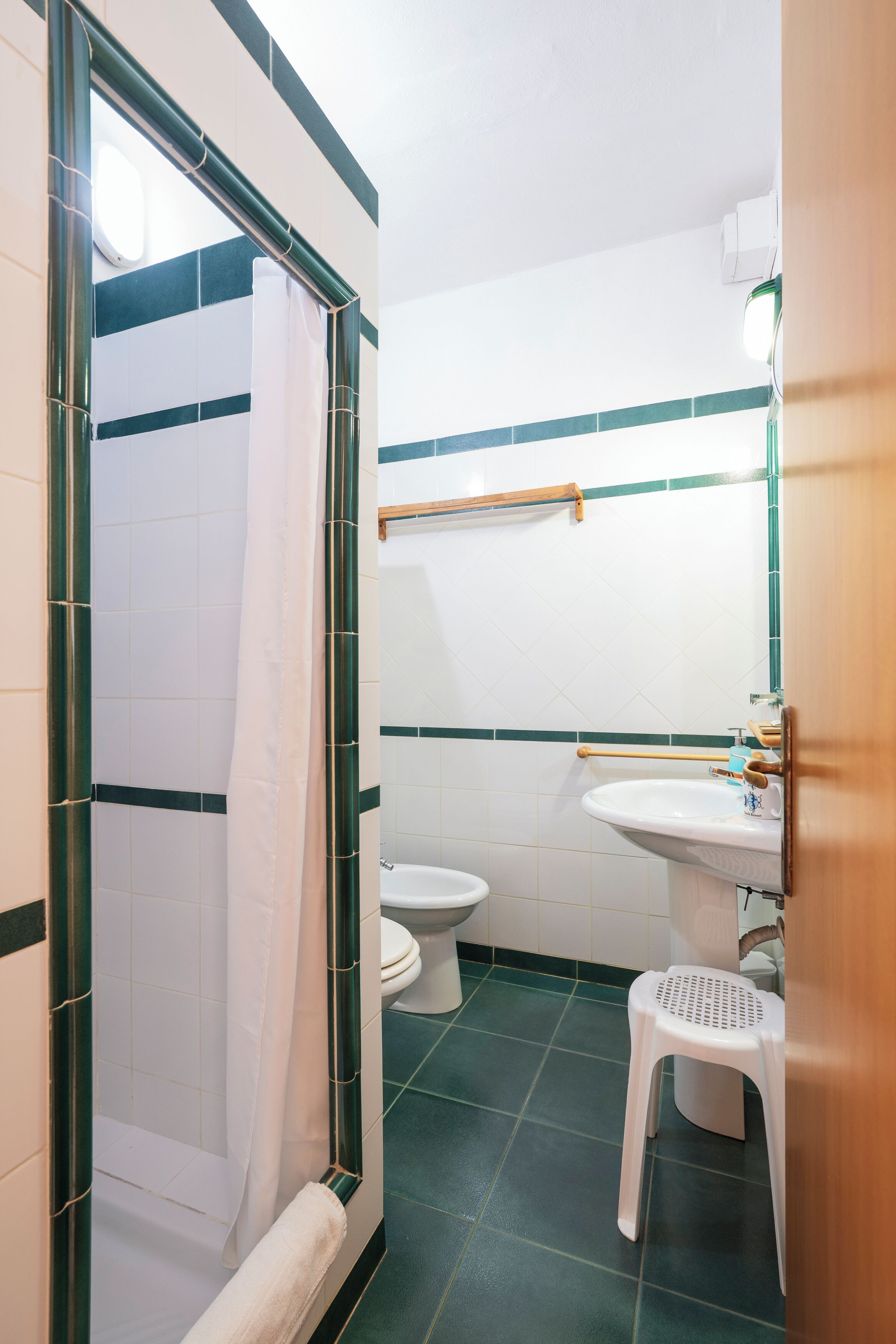 A bathroom with a shower, toilet, sink, and a small stool.