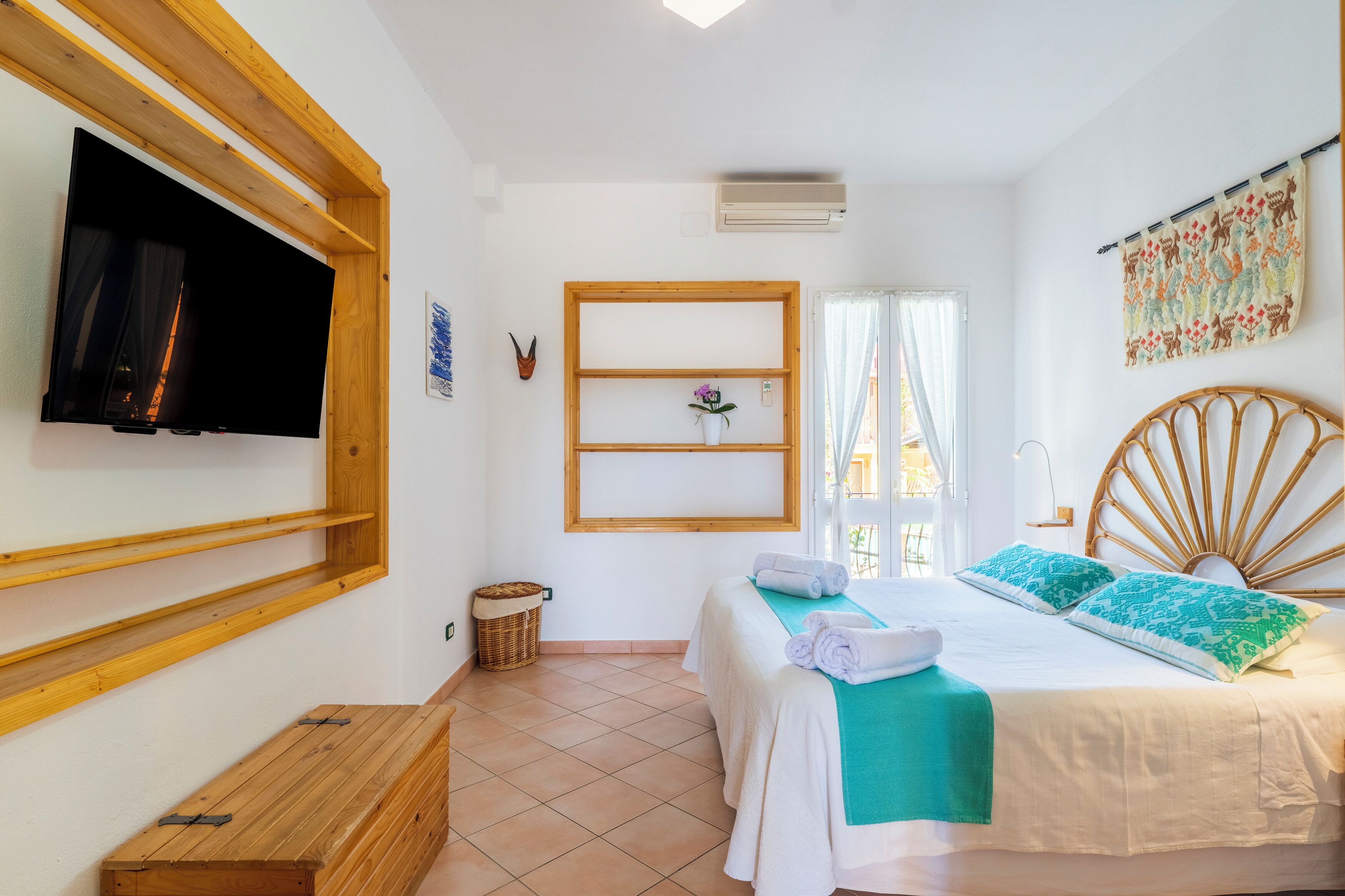 Une chambre à coucher avec un lit, un téléviseur fixé au mur, un banc en bois, une petite étagère et une fenêtre avec des rideaux.