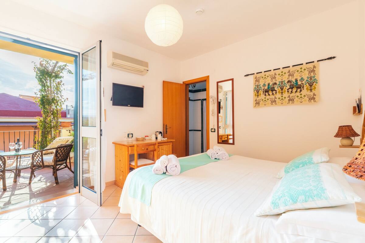 A hotel room with a bed, a desk, a chair, a TV, and a balcony with a table and chairs.