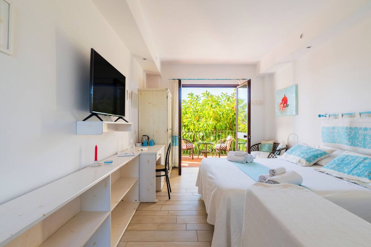 A hotel room with a bed, a desk, a chair, a TV, and a balcony with a view of greenery.